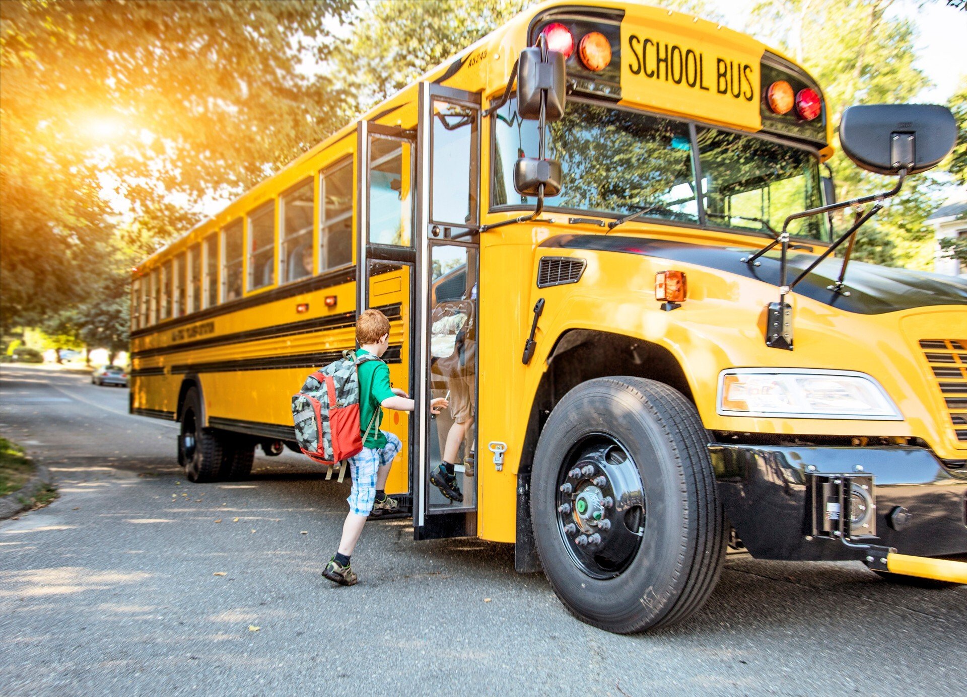 Young children with backpacks step onto a schoolbus with the sun shining through the trees in the background