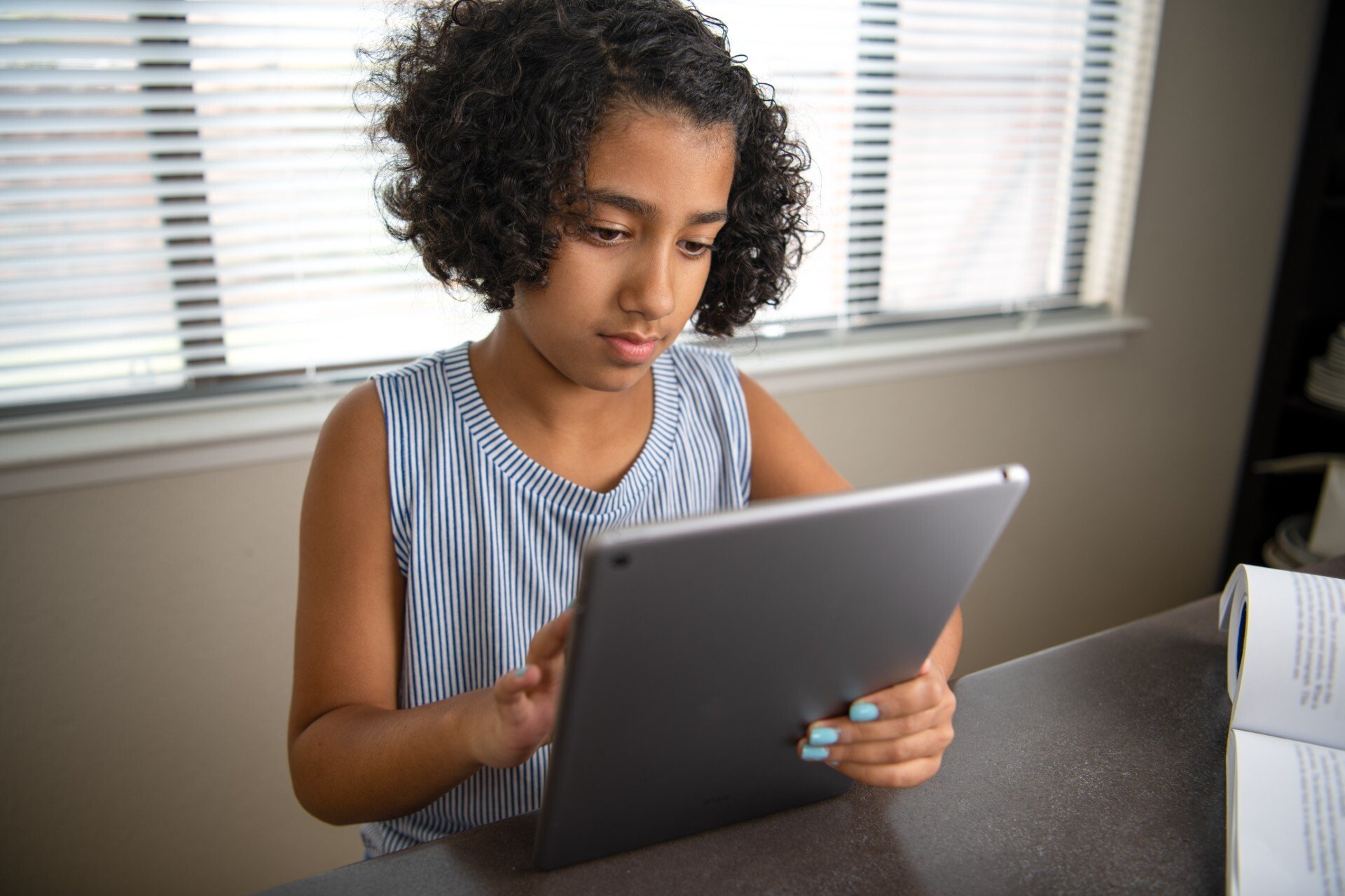 A school aged girl sits at a table looking at her tablet with an open book next to her