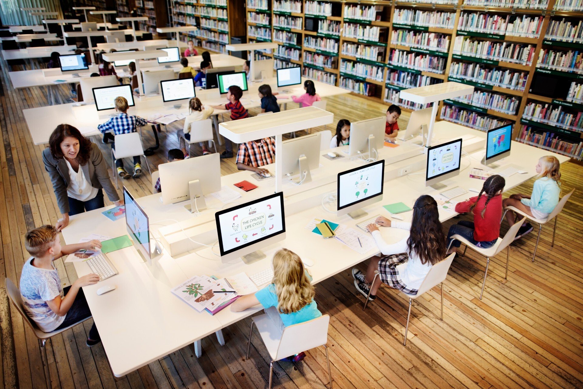A group of kids in a school library study at their computers with a librarian nearby to help them