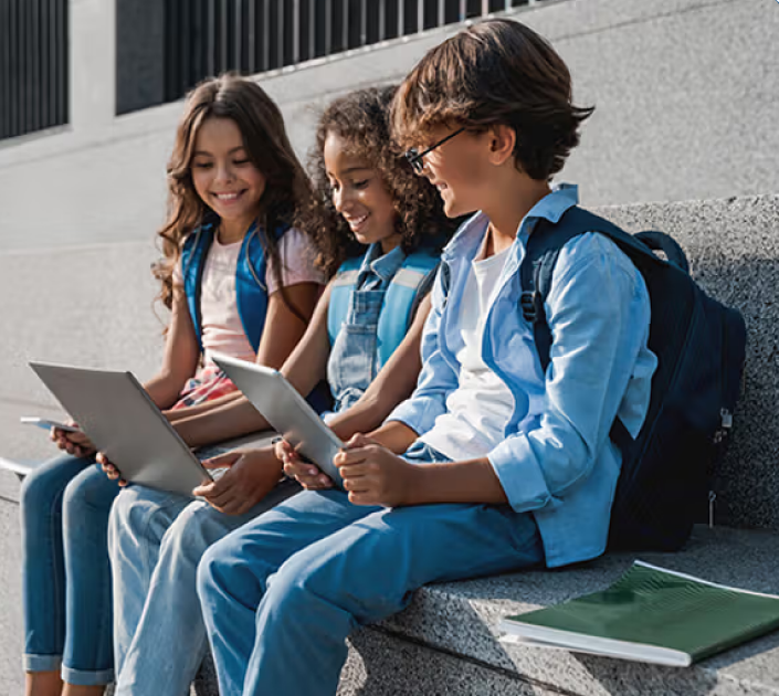 A group of smiling children sit on a stone bench using their tablets and smiling
