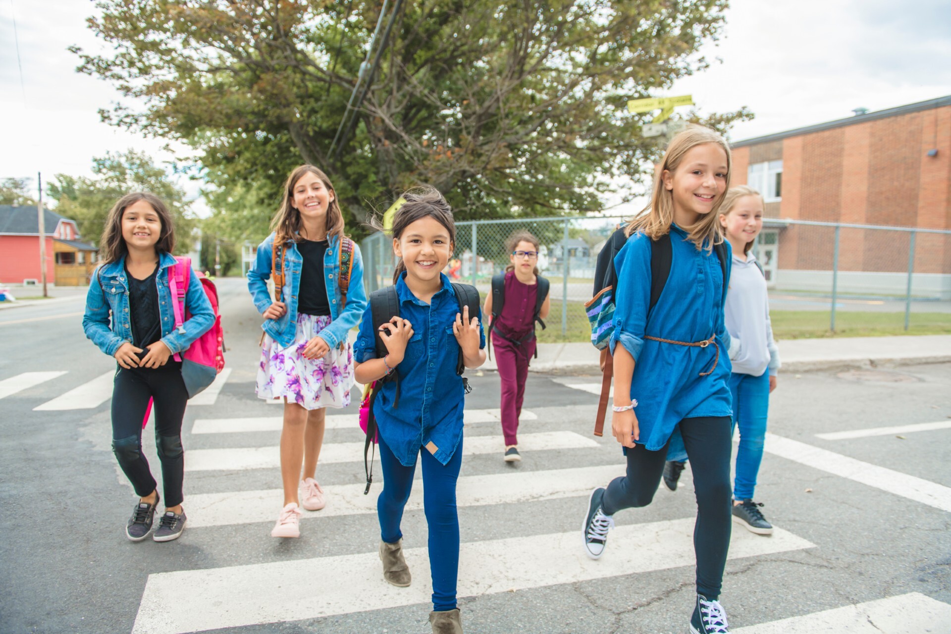 A group of kids wearing backpacks cross the street, smiling and looking at the camera